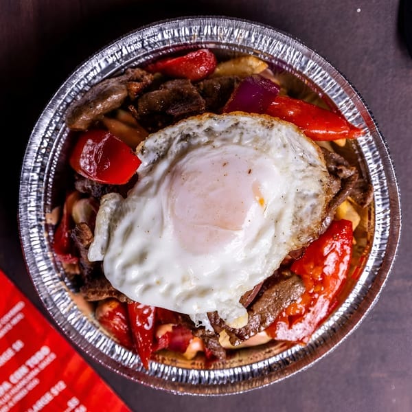 Peruvian Lomo Saltado in a foil container, topped with a fried egg, red peppers, and onions.