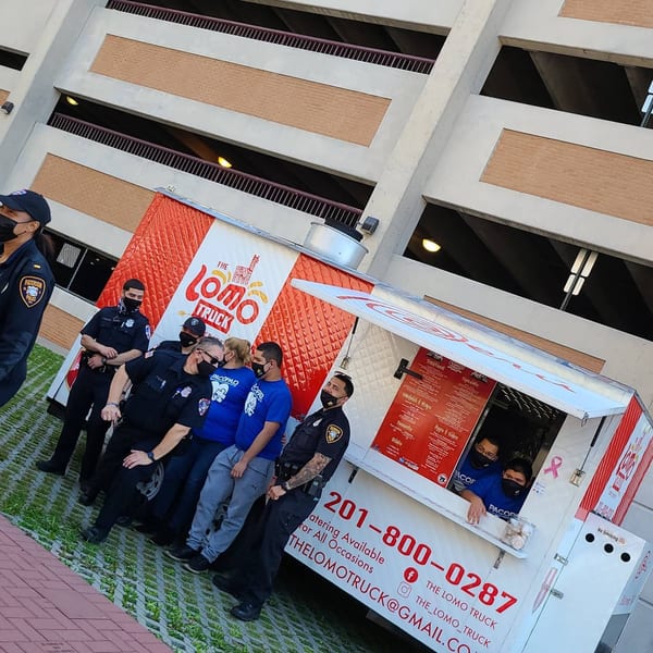 Police officers and staff stand by The Lomo Truck, a Peruvian food truck, with a parking garage in the background.