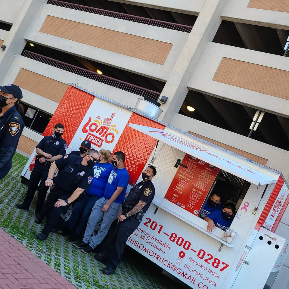 Police officers and staff stand by The Lomo Truck, a Peruvian food truck, with a parking garage in the background.