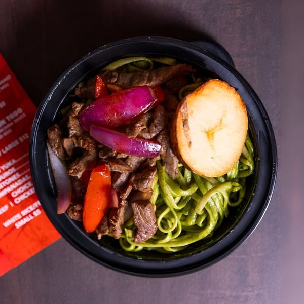 Peruvian Lomo Saltado with green noodles, red onions, bell peppers, and a fried plantain slice.