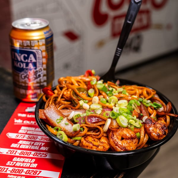 Peruvian Lomo Saltado with shrimp, noodles, and vegetables in a black bowl, with an Inca Kola can.