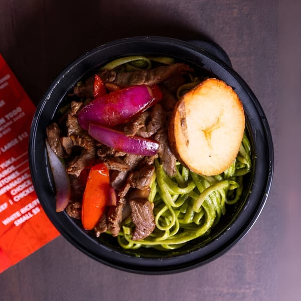 Peruvian Lomo Saltado with green noodles, beef, red onions, peppers, and a fried plantain slice.