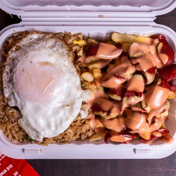 Peruvian chaufa with a fried egg and salchipapa with pink sauce in a white takeout container.